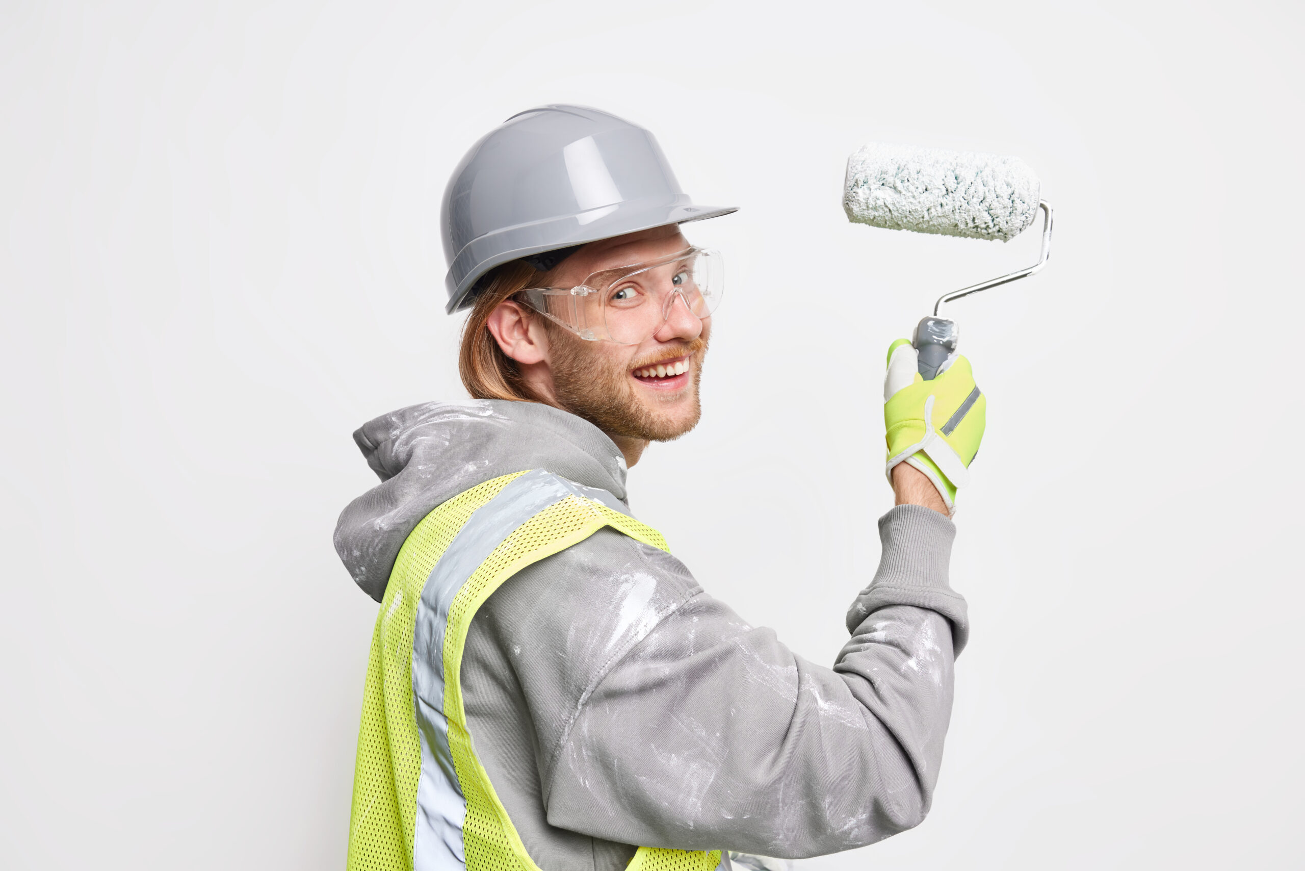 professional handyman paints new home holds roller wears protective helmet glasses and uniform does repair poses against white background. male repairr busy doing house renovation or interior renewal